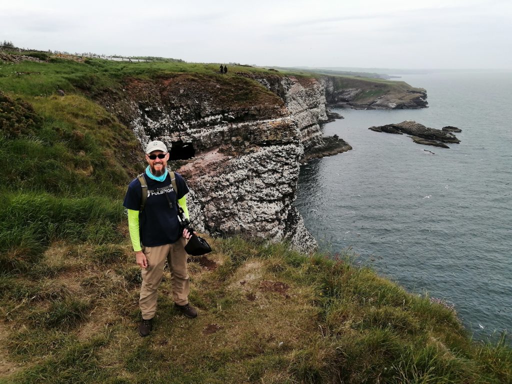 Posed photo of me standing near the edge of the Fowlsheugh cliffs. I am wearing a shirt with the Fulbright logo, sunglasses, & a baseball cap. I have a backpack on & am carrying my mic with its stand under my arm. The ground beneath my feet is rough & grassy. Beyond the edge behind me are several cliff faces completely covered in nesting sea birds. The water of the North Sea is 70 meters below. The sky is hazy.