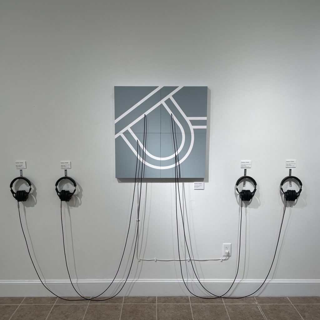 The complete installation. A flat sculpture hangs on the wall. Its surface depicts a map of the DeLeon Springs boil using simple geometric shapes made by white lines against a grey background. There are two headphones hanging from the wall on both sides of the sculpture. At four points on the map, cables emerge and droop toward the floor before reaching back up to four sets of headphones. 